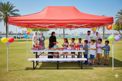 Children and adults gathered under a red tent with balloons and a picnic table on a sunny day.